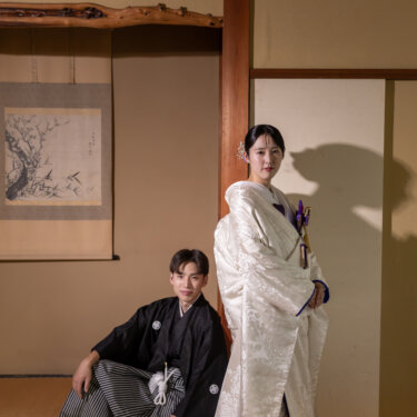 掛け軸の床の間の前に立つ白無垢の花嫁と、その足元にしゃがむ紋付袴の新郎。小石川後楽園の和室で撮る結婚写真