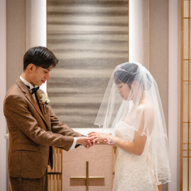 十字架のある祭壇の前で、ベールをかぶった花嫁の手を取り目を伏せるツイードスーツの新郎。誓いの場面を再現したチャペルフォト