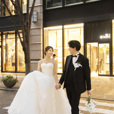 ガラス張りの店舗が並ぶ石畳を手つなぎで歩く、タキシードの新郎とウェディングドレスの花嫁。おしゃれな街ロケの前撮り