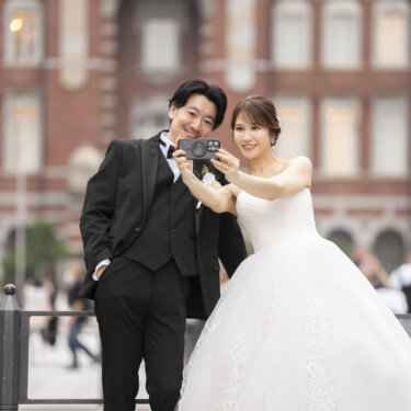 東京駅丸の内駅舎の前で、カメラを手に2人で自撮りをする新郎とウェディングドレスの花嫁。楽しい雰囲気の洋装前撮り