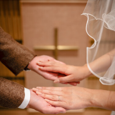 十字架の祭壇を背に、新郎の手のひらに花嫁が手を重ねる手元のアップ。結婚指輪が光るチャペルフォトの一枚