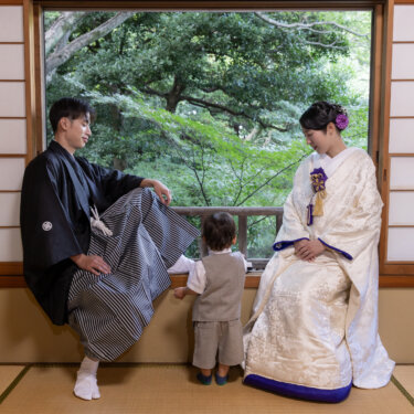 緑あふれる庭を望む窓辺に座る紋付袴の新郎と白無垢の花嫁、その間で外をのぞく男の子。家族で撮る和装前撮り