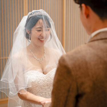 新郎の肩越しに見た、ベール越しに笑うティアラの花嫁。レースのウェディングドレスで撮るチャペルでの前撮りのワンシーン
