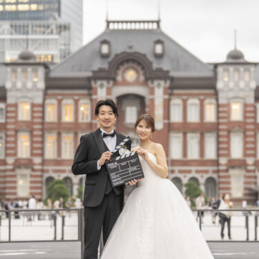 東京駅丸の内駅舎のドームを背に、カチンコを持って並ぶ新郎と花嫁。映画風の小物を使った洋装フォトウェディング