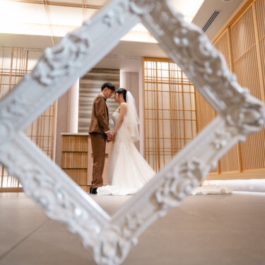 白いアンティークの額縁越しに、チャペルでおでこを寄せ合う新郎新婦を写した構図。額縁フレームを前ボケに使った結婚写真