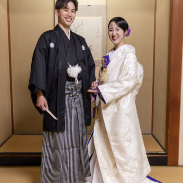 床の間の前で寄り添い、カメラに笑顔を向ける紋付袴の新郎と白無垢の花嫁。和室で撮る全身の結婚写真