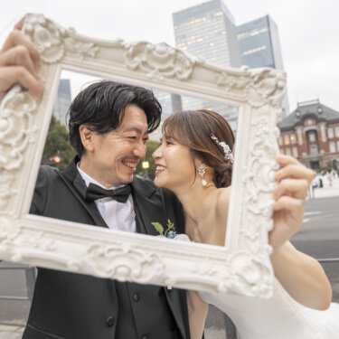 白い額縁を2人で持ち、フレームの中で頬を寄せて笑う新郎とウェディングドレスの花嫁。東京駅前で撮る小物アイデアの前撮り