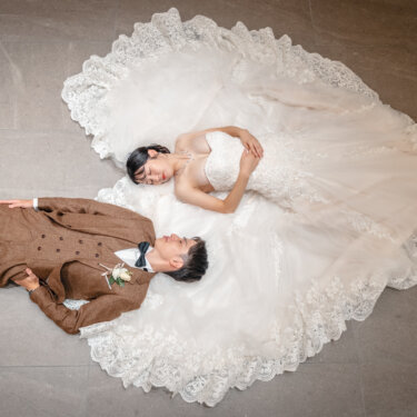 床いっぱいに広げたレースのトレーンの上で、頭を寄せて横たわる新郎新婦を真上から撮影したウェディングフォト