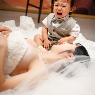 寝転ぶウェディングドレスの花嫁のそばで、泣き顔を見せるベスト姿の男の子。子連れの前撮りならではの自然な一コマ