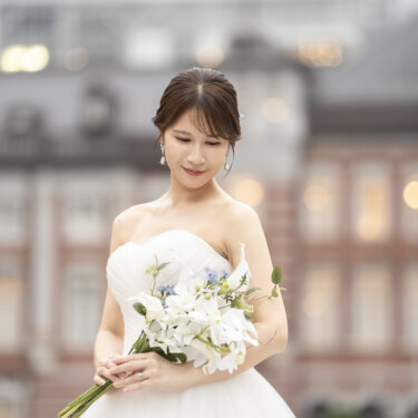 赤レンガの建物をぼかした背景に、白いブーケを持ってうつむくウェディングドレスの花嫁。プレ花嫁のソロカットの前撮り