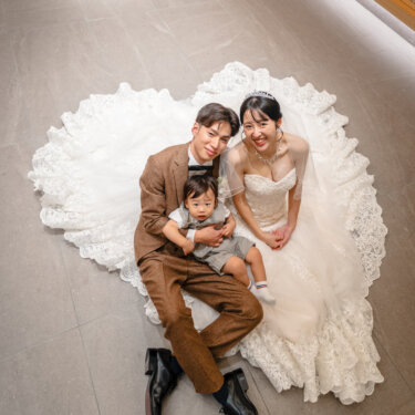 円く広げたレースのトレーンの上に座り、息子を抱いて見上げる新郎新婦。チャペルで撮る家族写真のフォトウェディング