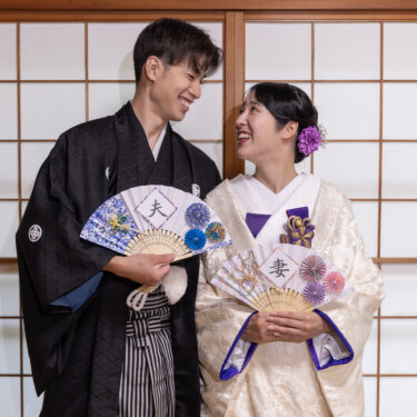'夫''妻'の文字入り扇子を手に、障子の前で見つめ合って笑う紋付袴の新郎と白無垢の花嫁の和装前撮り