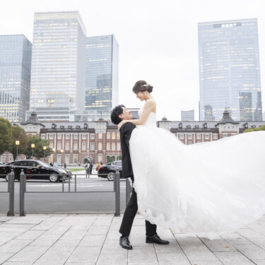 高層ビルと駅舎を背に、ウェディングドレスの裾をなびかせて花嫁を抱き上げる新郎。丸の内の広場で撮る動きのある前撮り