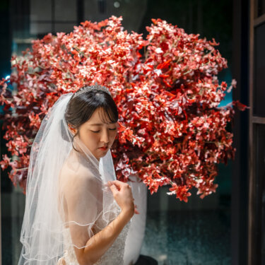 赤い花の大きな装花を背に、ティアラとベールの花嫁が伏し目がちに佇むブライダルポートレート。ウェディングドレスの洋装前撮り