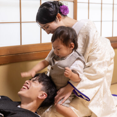 畳に寝転んで笑う新郎の顔に手を伸ばす赤ちゃんと、その子を抱いてほほえむ白無垢の花嫁。家族で撮る結婚写真