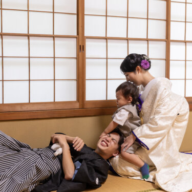 和室の畳に寝転ぶ紋付袴の新郎に、白無垢の母に支えられた赤ちゃんがじゃれつく家族写真。にぎやかな和装前撮り
