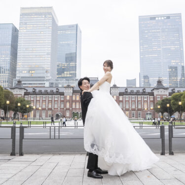 高層ビルの前で、ウェディングドレスの花嫁を抱き上げてくるりと回る新郎。広場で撮る動きのあるフォトウェディング