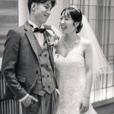 新郎の肩に腕をかけて大笑いするベールの花嫁と、ポケットに手を入れて微笑む蝶ネクタイの新郎。モノクロで残すナチュラルな結婚写真