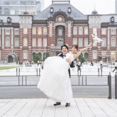 東京駅丸の内駅舎の正面で、ブーケを掲げた花嫁をお姫様抱っこする新郎。開放感のある洋装前撮りのポーズ