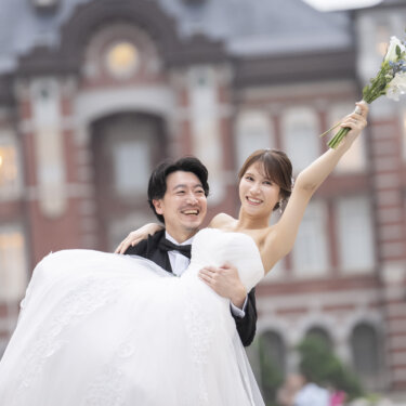 赤レンガの駅舎の前でお姫様抱っこされ、ブーケを高く掲げて笑うウェディングドレスの花嫁。笑顔あふれる結婚写真