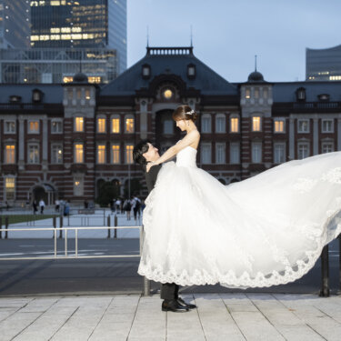 夕方の駅舎を背景に、ドレスの裾を大きく広げて花嫁を抱き上げる新郎。ライトアップ前の時間に撮るウェディングフォト