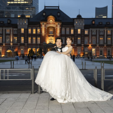 ライトアップが始まった東京駅丸の内駅舎の前で、ウェディングドレスの花嫁をお姫様抱っこする新郎。夜景の前撮り