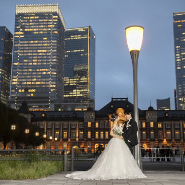 高層ビルの夜景と街灯の下で寄り添う、ウェディングドレスの花嫁とタキシードの新郎。夜に撮るロマンチックな前撮り