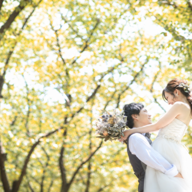 黄色く色づいた木々の下で、ドライフラワーブーケを持つウェディングドレスの花嫁を抱き上げる新郎。秋の洋装前撮り