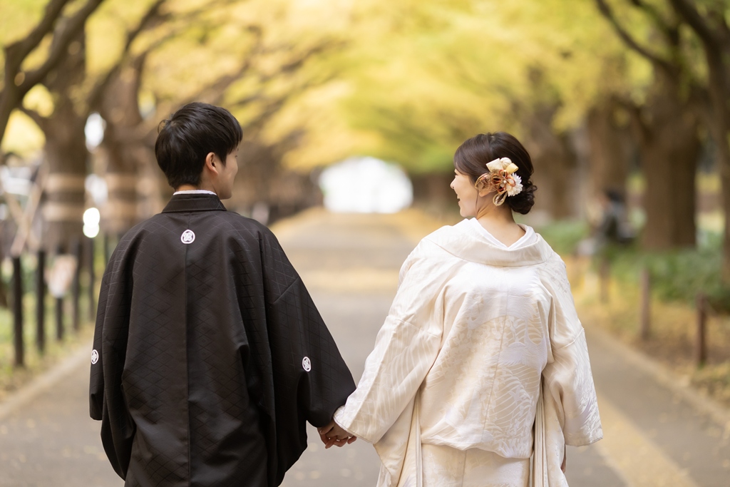 外苑いちょう並木の歩道を、手をつないで歩く白無垢の花嫁と紋付袴の新郎の後ろ姿