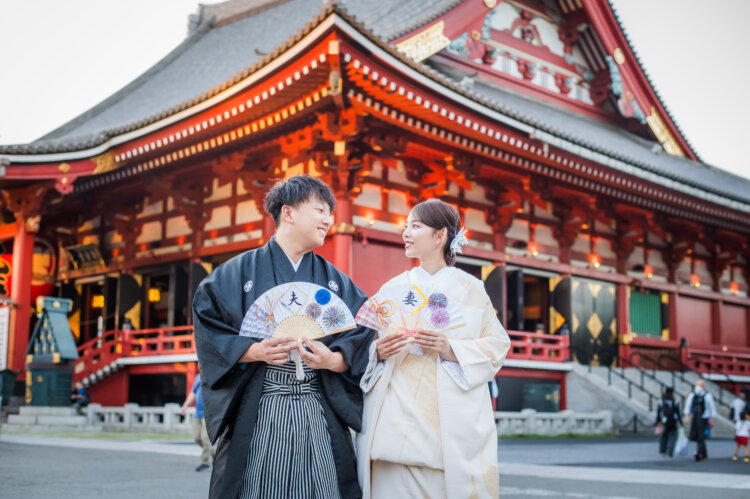 浅草寺の朱色の本堂を背に、'大''吉'の文字入り扇子を手に寄り添う紋付袴の新郎と白無垢の花嫁の和装前撮り