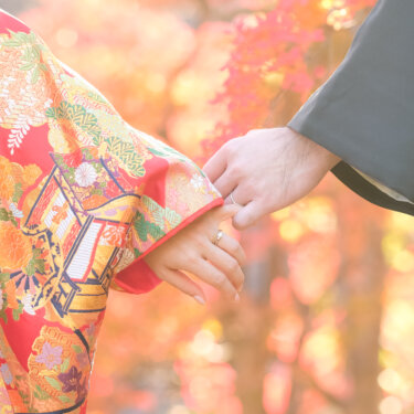紅葉の光を背に手を取り合う、赤い色打掛の花嫁と紋付袴の新郎の手元。和装前撮りで人気の指輪ショット