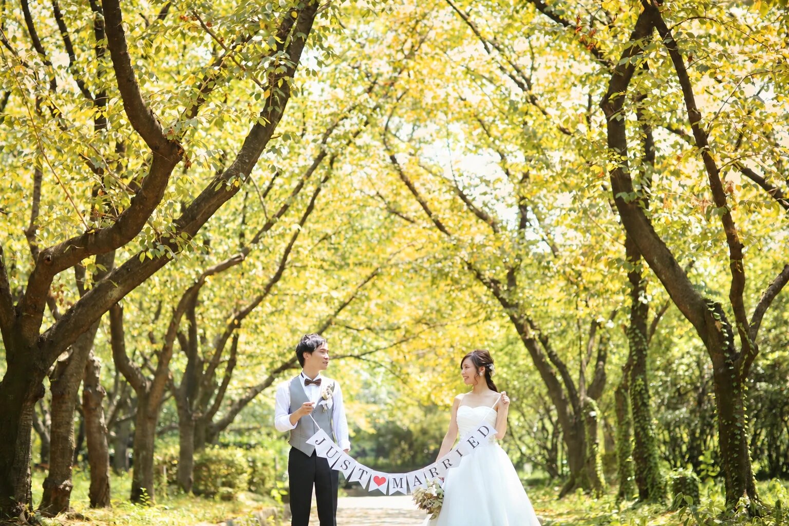 黄色く色づいた並木道で「JUST MARRIED」のガーランドを持って向かい合うベスト姿の新郎とウェディングドレスの花嫁。秋の洋装前撮り
